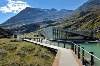 View of the Trollstigen Visitor Centre and a path by a river surrounded by greenery and mountains under a blue cloudy sky