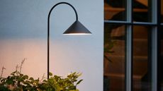 Image of a black garden stake light illuminated against a white outdoor wall with plants around the bottom of it.