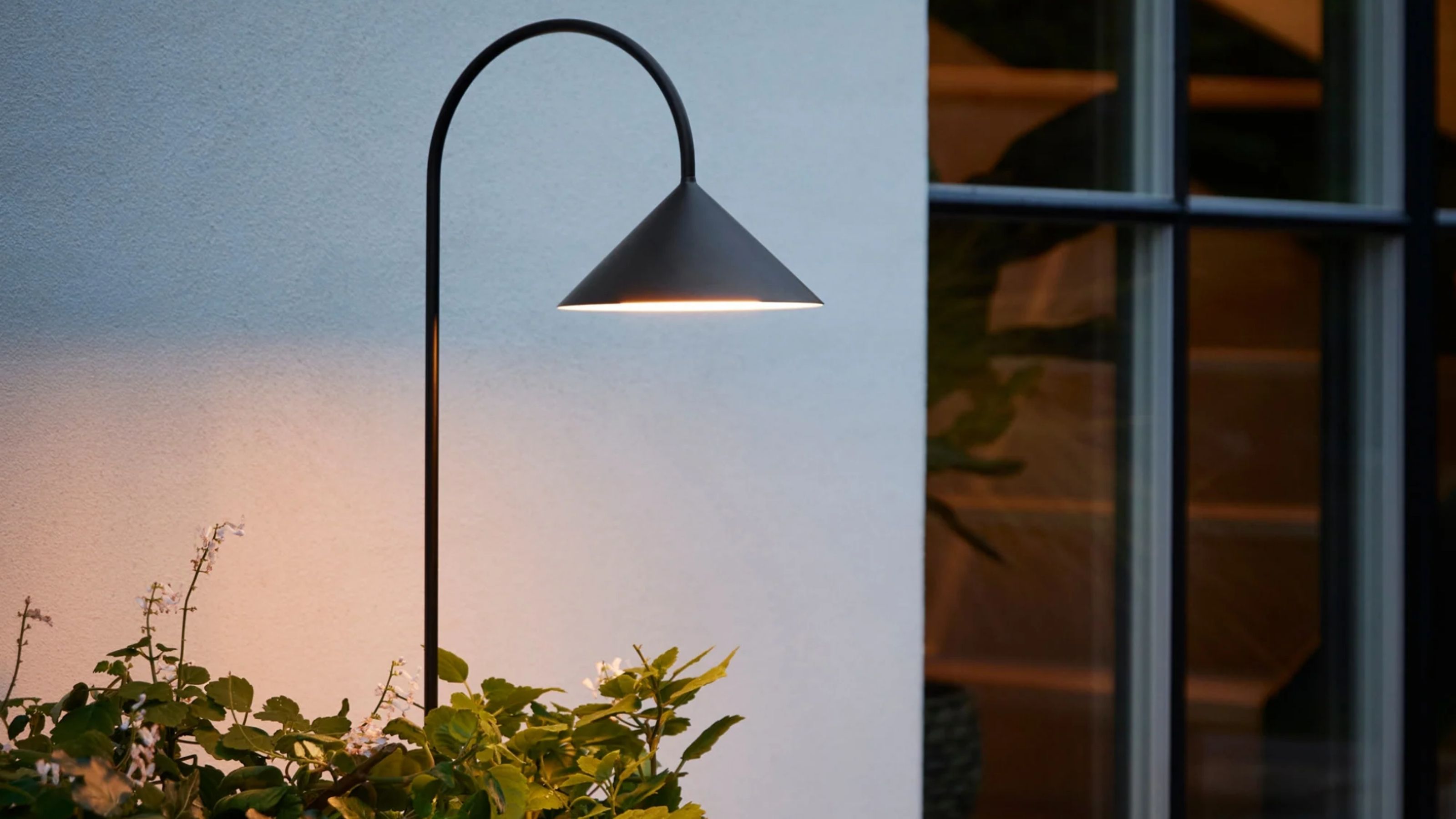Image of a black garden stake light illuminated against a white outdoor wall with plants around the bottom of it.