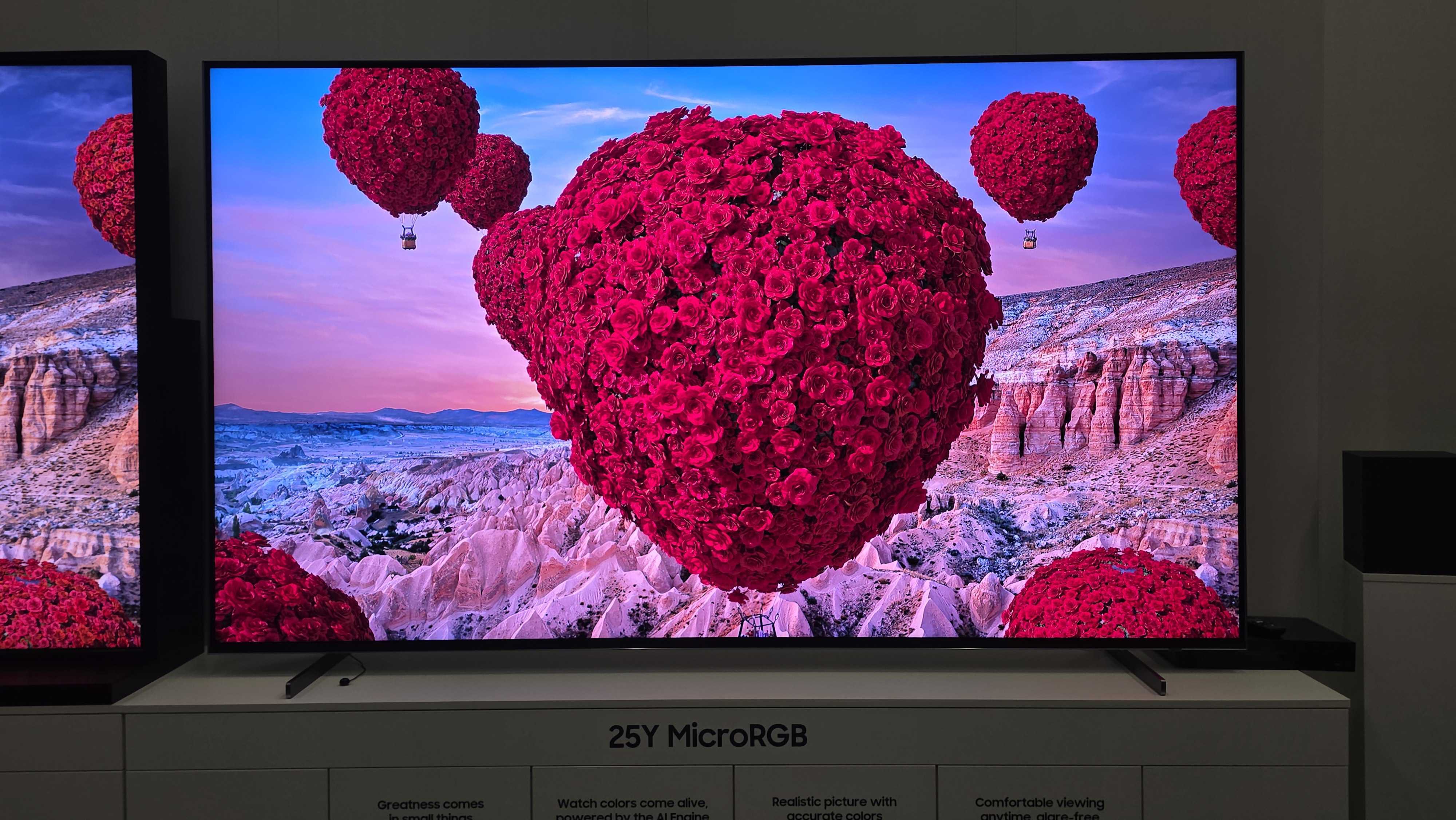 A 115-inch Samsung RGB LED TV on a white stand with colourful demo content on screen featuring the Statue of Liberty and a hot air balloon made of roses.