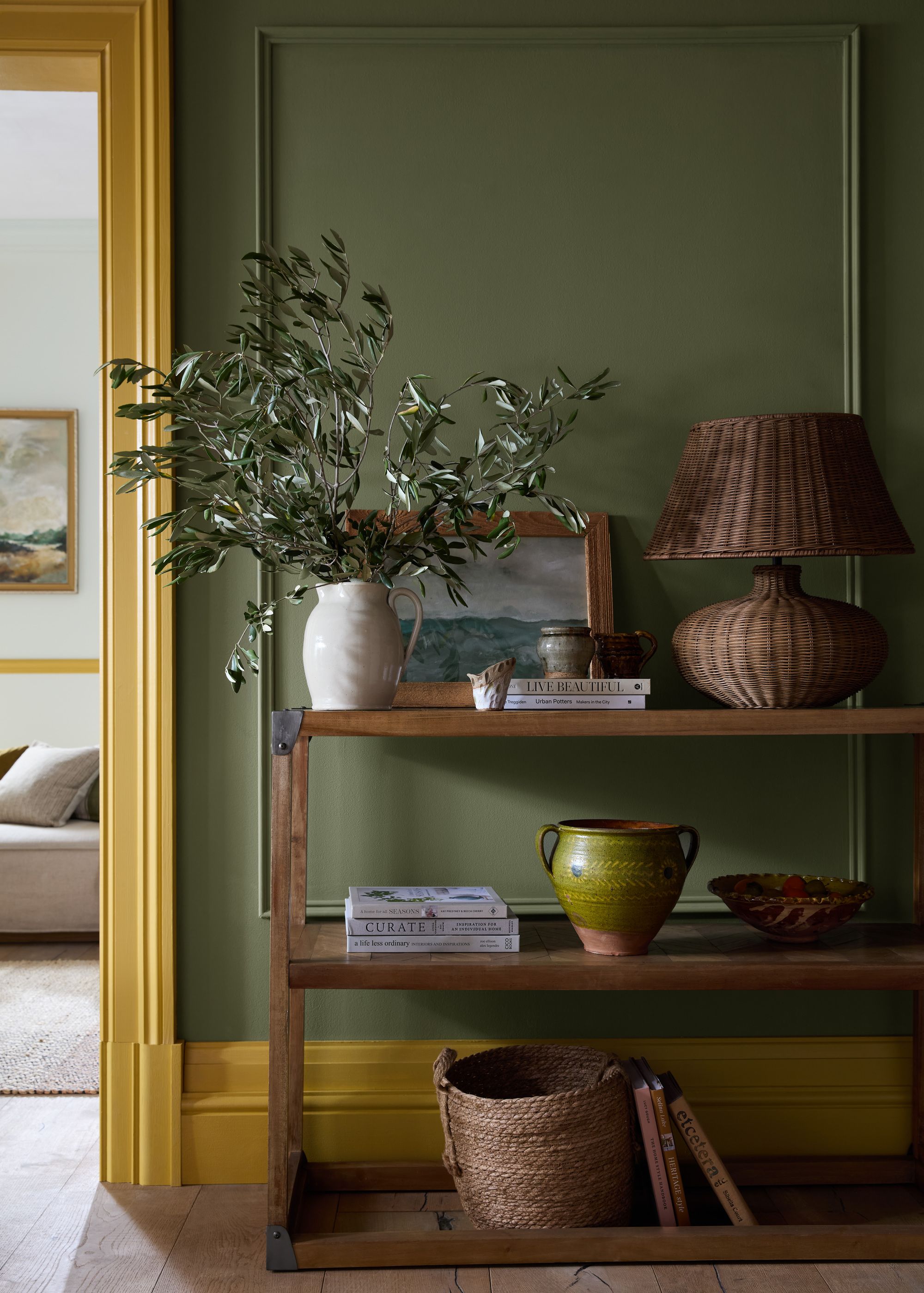 Image of an olive green wall in a hallway with golden yellow trim. There is a wooden console with three shelves. On the shelves is a wicker lamp, a white vase with green stems, coffee table books, and a few vases and catchalls.