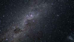 The glowing band of the Milky Way can be seen streaking across an image captured from near lunar space, from the bottom left to top right of the photo. Pink nebulas and dark dusty clouds can be seen threading the galactic plane, and a satellite galaxy is visible as a smudge of white light to the bottom right of the screen.