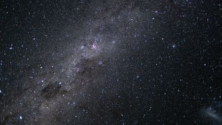 The glowing band of the Milky Way can be seen streaking across an image captured from near lunar space, from the bottom left to top right of the photo. Pink nebulas and dark dusty clouds can be seen threading the galactic plane, and a satellite galaxy is visible as a smudge of white light to the bottom right of the screen.