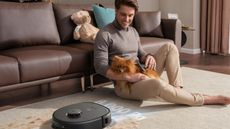 A man sitting on a cream carpeted floor holding a small tan dog and a robot vacuum cleaning around them