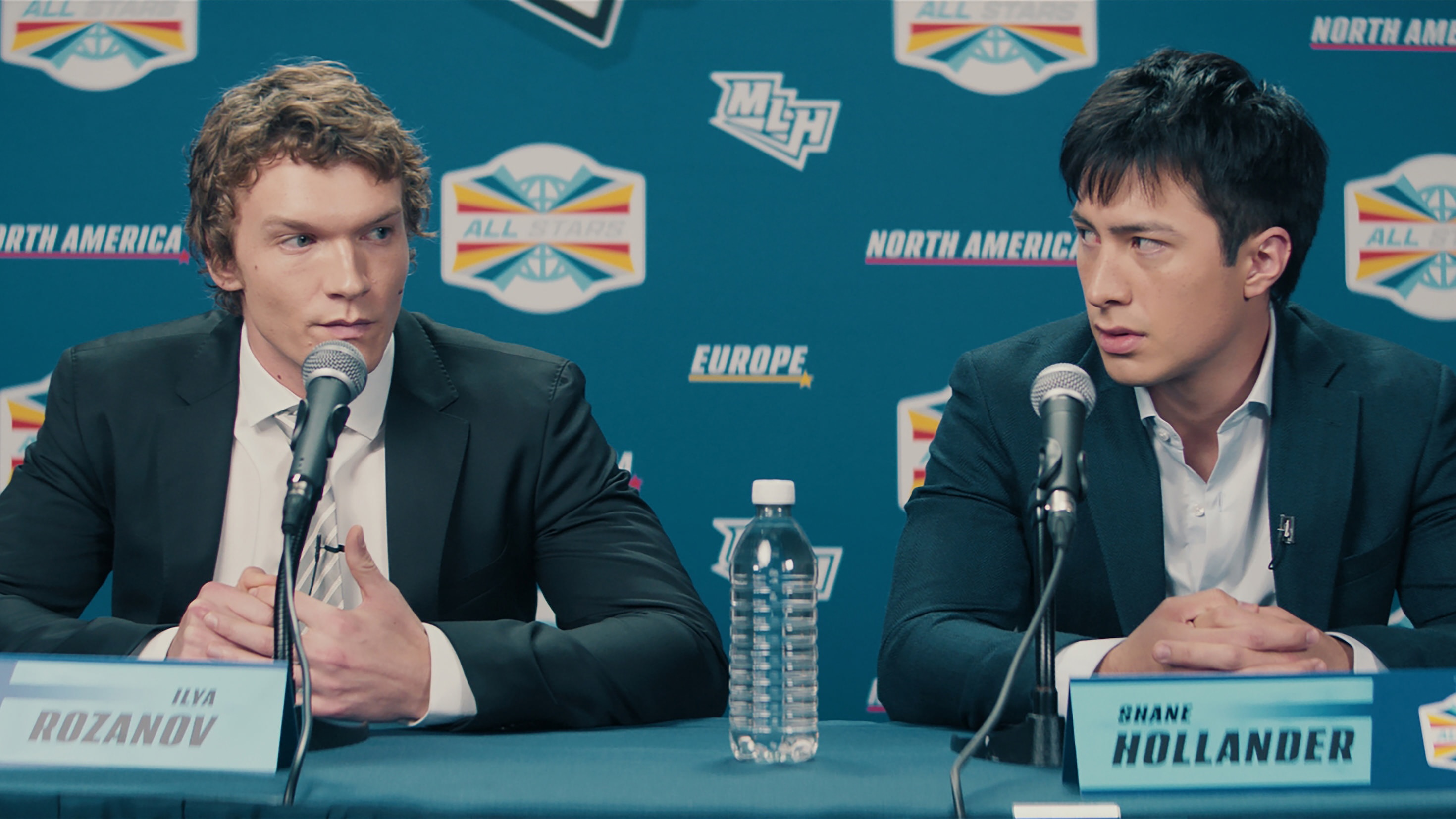 Two men (Connor Storrie as Ilya Rozanov and Hudson Williams as Shane Hollander) sit in front of mics and name plates at a press-conference table, in the TV show &#039;Heated Rivalry.&#039;