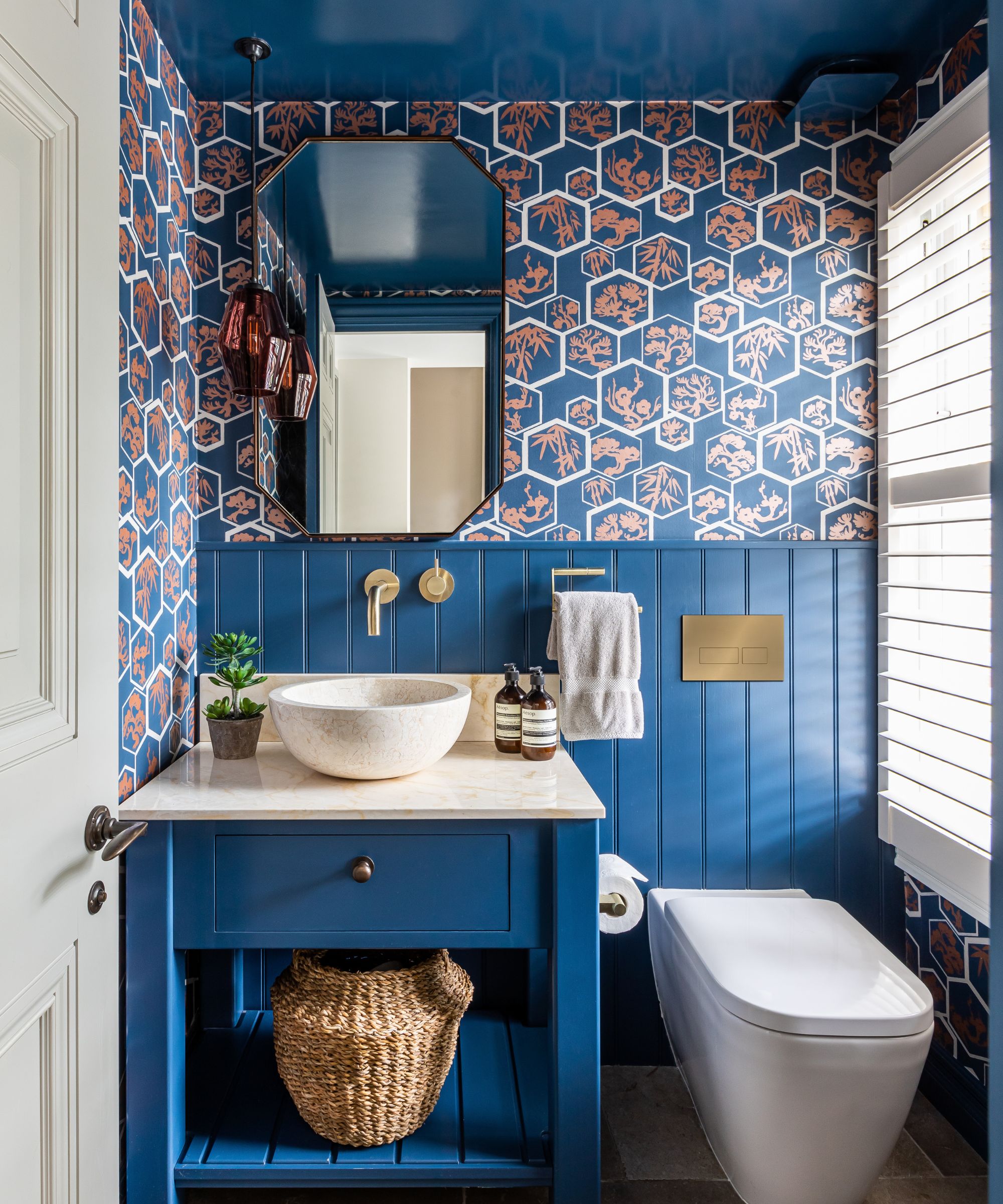 Bathroom with blue ceiling and blue painted wood panelling, blue vanity unit with marble countertop and sink, wallpaper on top half of wall, long window with white blind and gold hardware