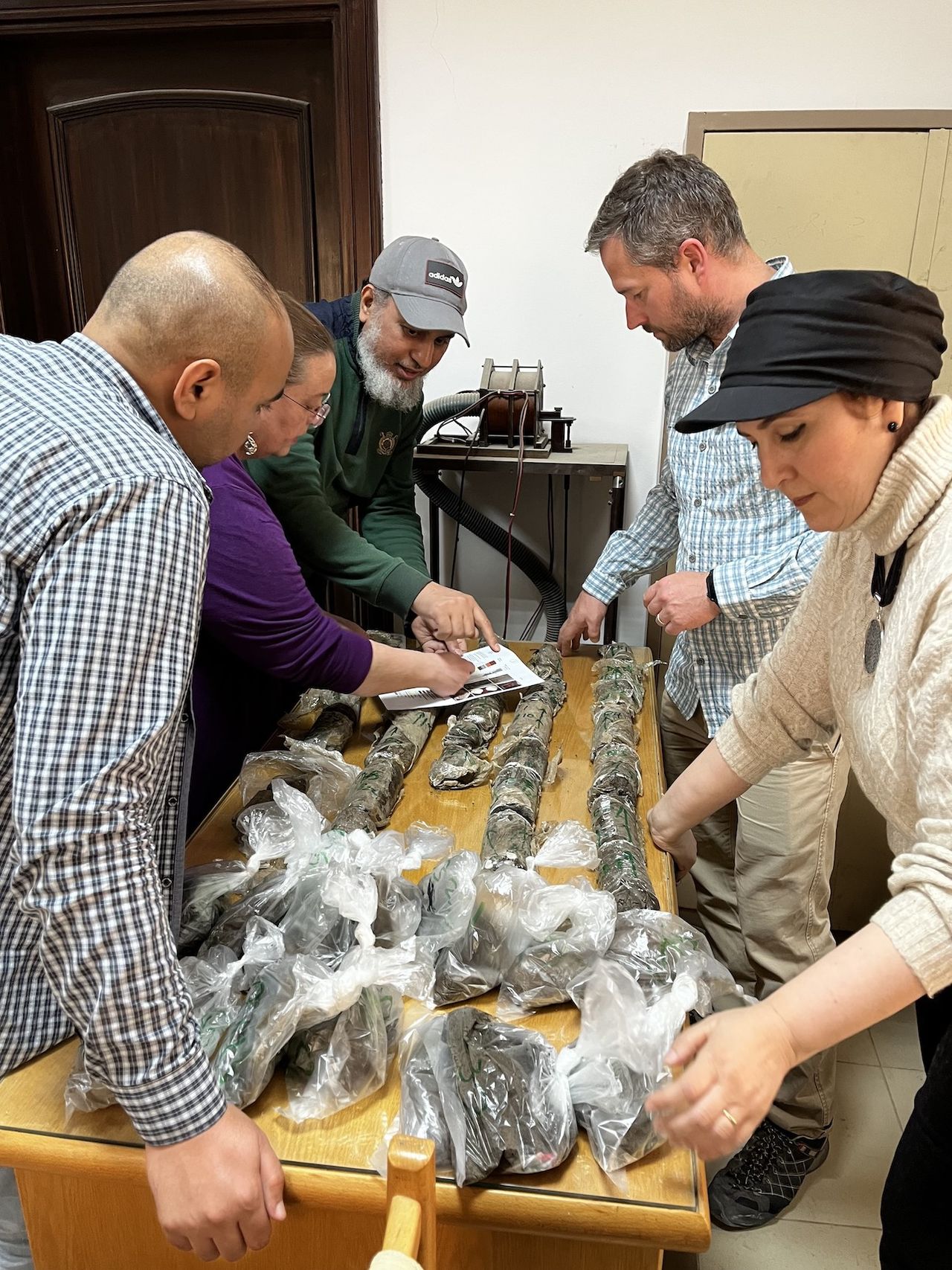 Five people stand around a table that has bags of soil samples on top of it. One man and woman both point to a piece of paper on top of some of the samples with photographs printed on it