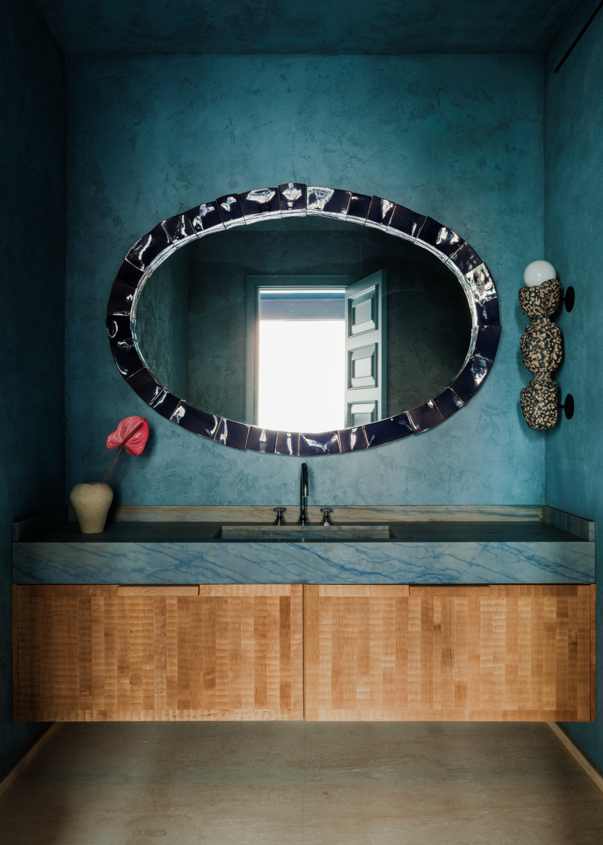 A bathroom with textured blue paint, a statement mirror, a speckled wall sconce, a ceramic with an anthurium, a blue marble counter, and wood cabinets