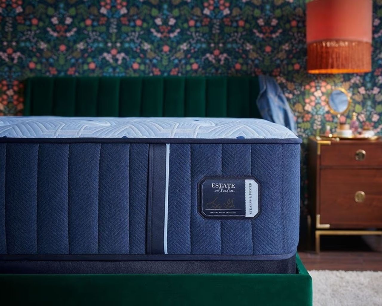 A blue Stearns &amp; Foster estate mattress on a green velvet bed, with statement green and blue wallpaper, and to the right of the bed is a wooden bedside table, with an orange lampshade above.