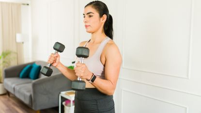 woman sideways to the camera holding two dumbbells in a hammer curl position. there's a sofa further away behind her and she's standing against a white wall.