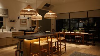kitchen at night with 3 large wicker pendants hung over black marble topped kitchen island with seating and storage
