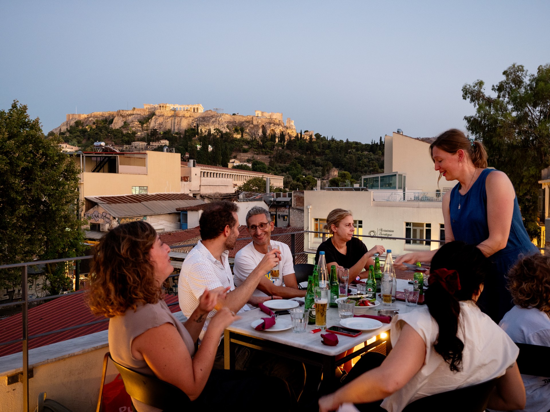 A group of friends share beers and food around a table on an Athens rooftop terrace at dusk, with the floodlit Acropolis rising on the hill behind them.