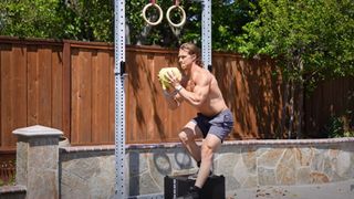 Man on a box holding a kettlebell with box hands performing a pistol squat in the garden