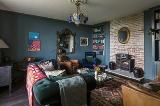 Living room or cosy snug with dark blue walls, brick chimney breast with lit woodburner. patterned cushions and eclectic mixture of antique furniture