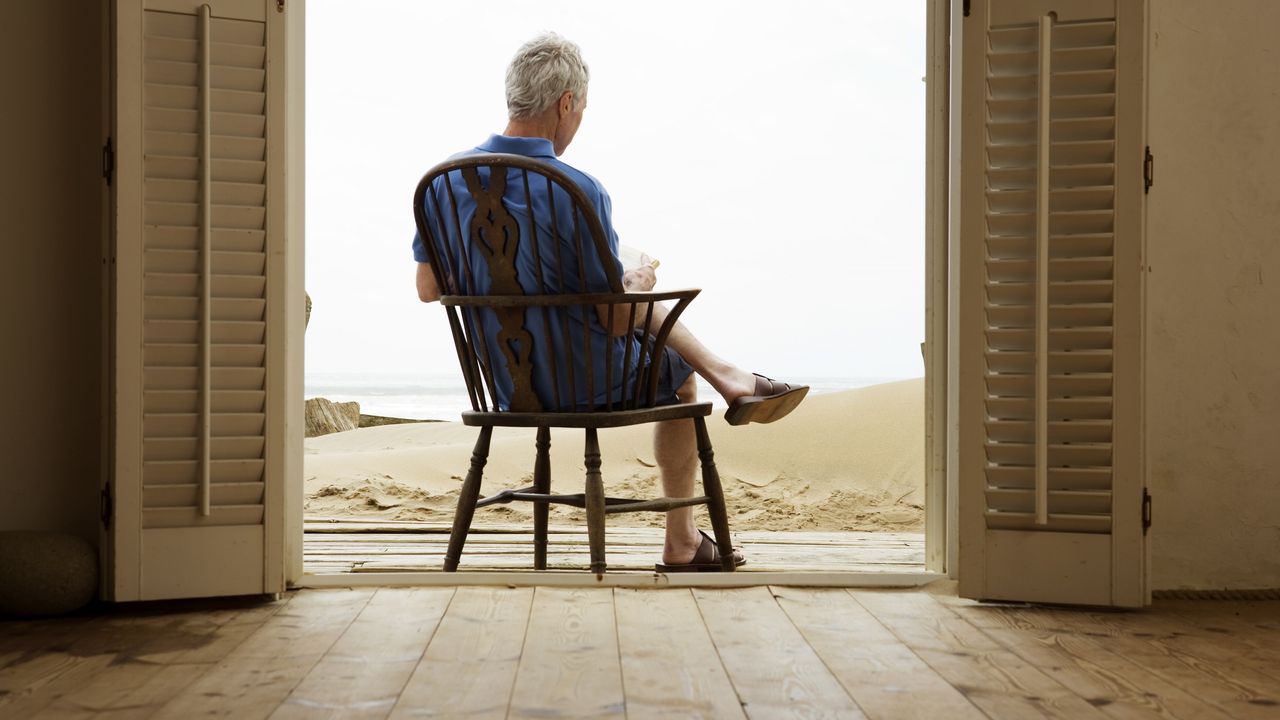 Older man who&#039;s reading sits in a chair facing the beach in the open double doorway of a beach house.