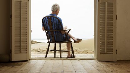 Older man who's reading sits in a chair facing the beach in the open double doorway of a beach house.