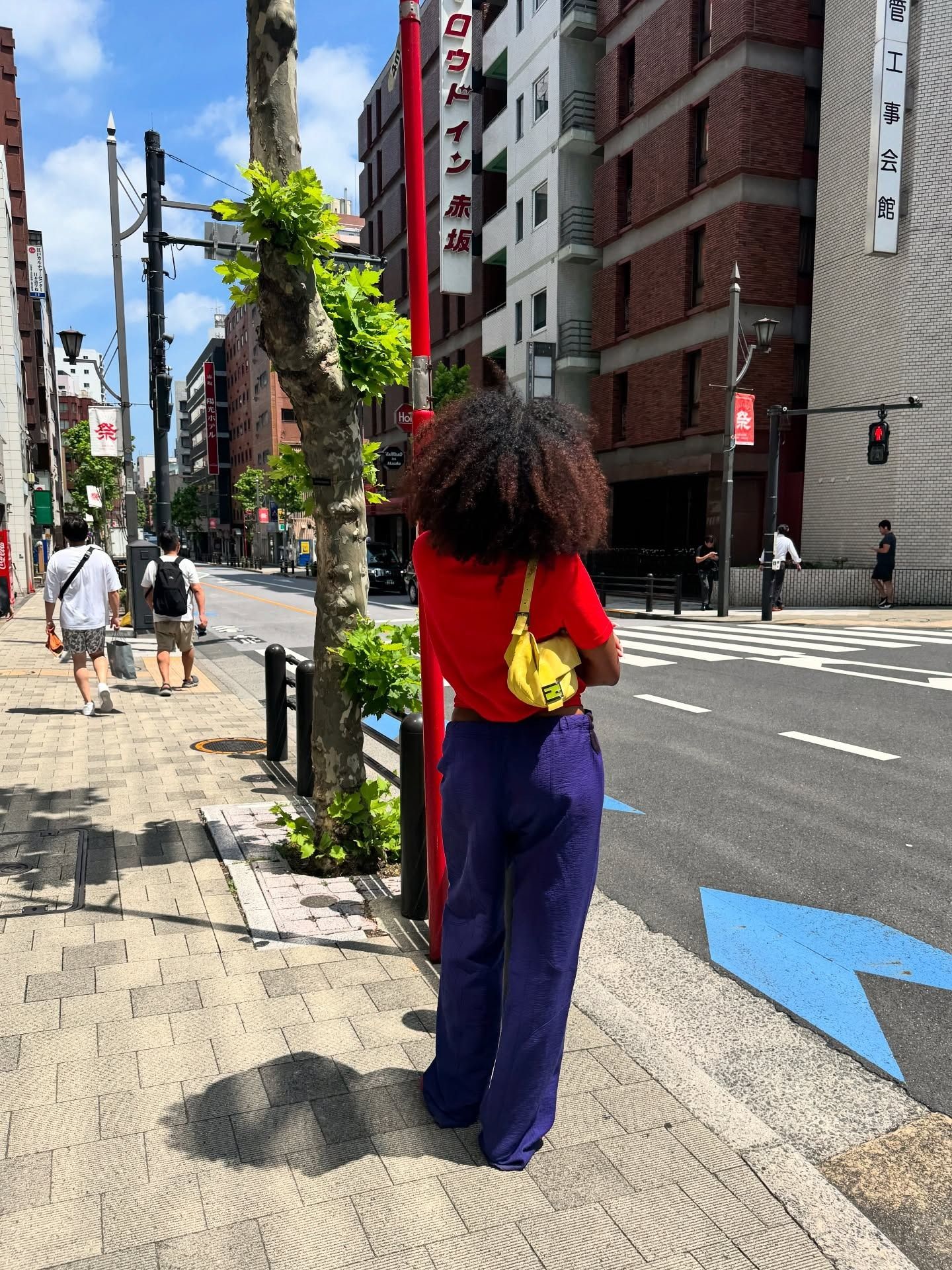 Naomi wears a red tee, yellow bag, and purple pants.
