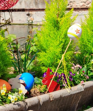 A garden planter with white and red bunny stakes in it, white, purple, and pink flowers, tall green shrubbery and a river in the background