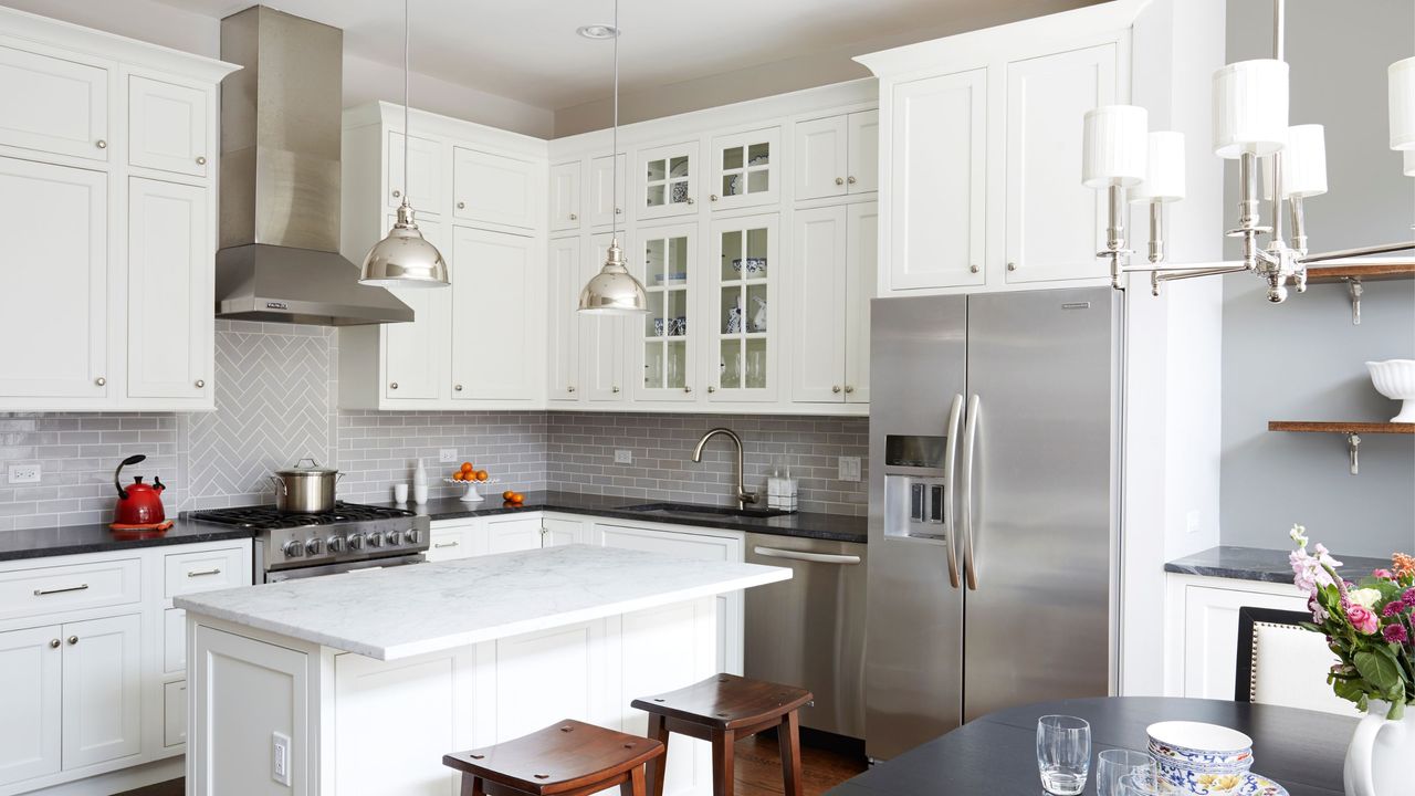 Narrow kitchen island ideas are so chic. Here is a kitchen with a white marble kitchen island with dark wooden stools in front of it, two silver pendant lights above it, and white cabinets and a silver oven behind it