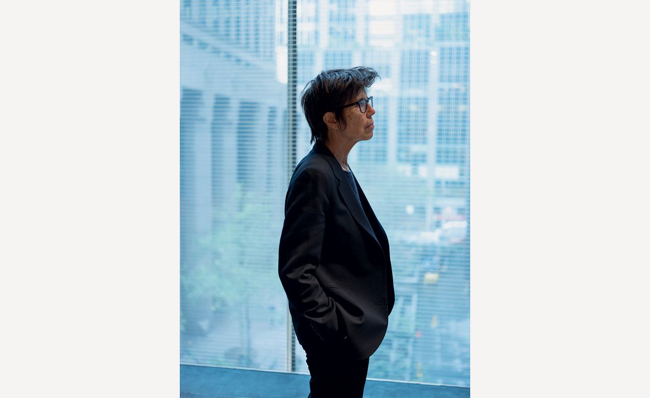 A woman with short hair stands side-on to the camera, wearing glasses and a black suit