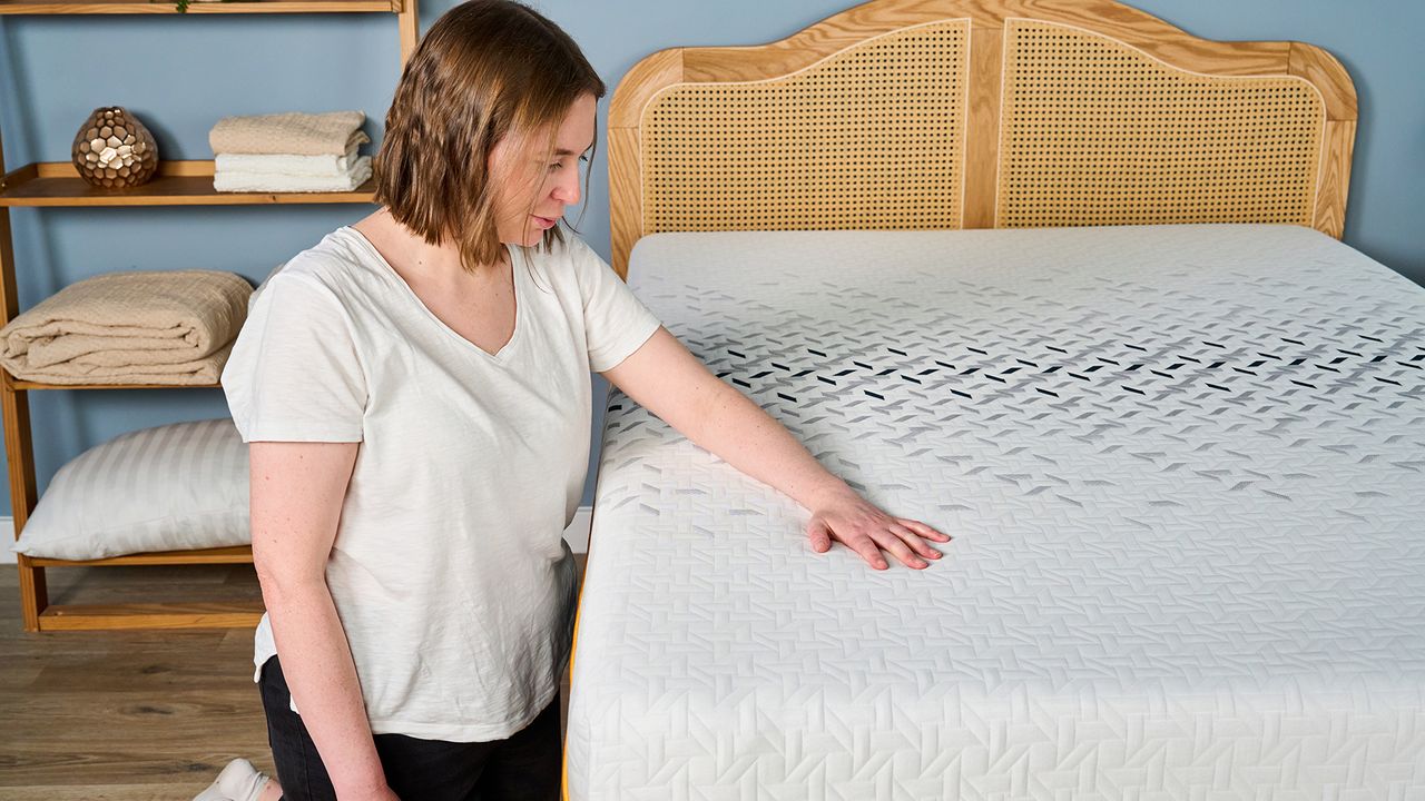 A female Tom's Guide tester kneeling next to the Nectar Ultra Hybrid mattress and feeling the soft cooling cover with her arm and hand