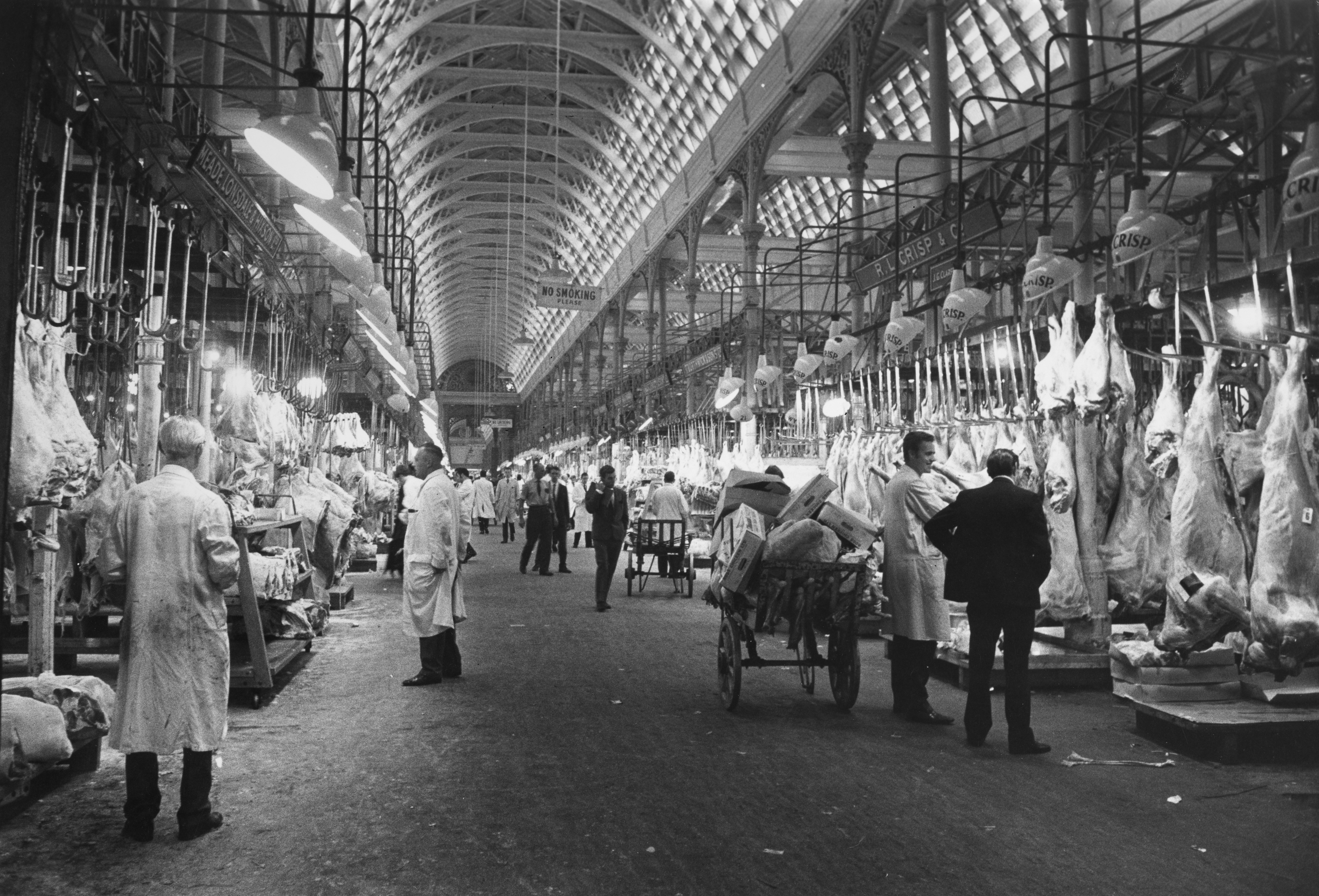 Rows of animal carcasses hang from hooks, beside market traders, porters and shoppers at Smithfield Meat Market