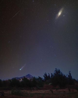 comet streaks across the sky and glows green, with a bright galaxy in the top left corner and a long thin meteor trail in the sky to the top left corner.