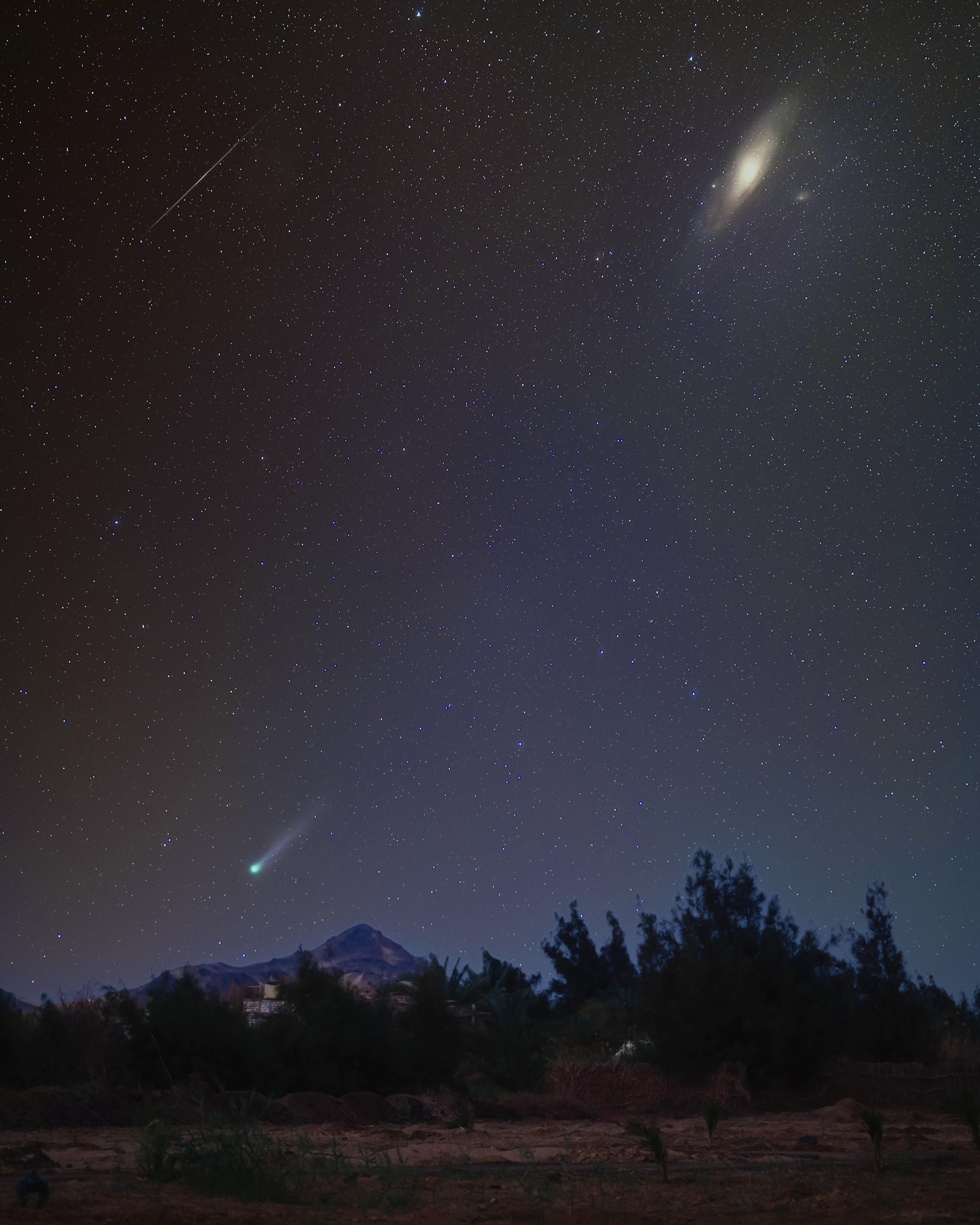 comet streaks across the sky and glows green, with a bright galaxy in the top left corner and a long thin meteor trail in the sky to the top left corner.
