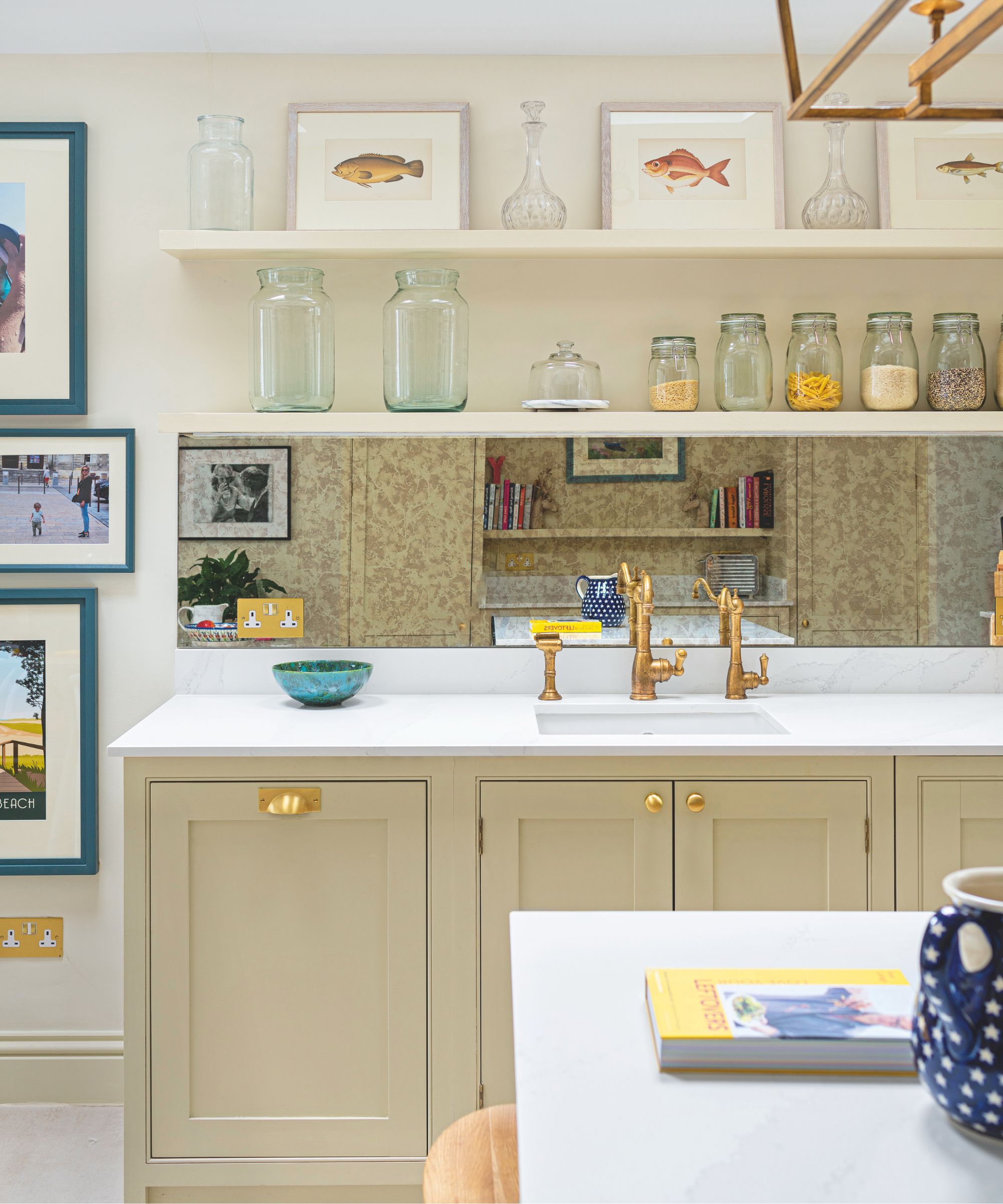 A cream kitchen with a mirrored backsplash, beige cabinets, gold hardware and faucets, white countertops, gallery wall and white kitchen table.
