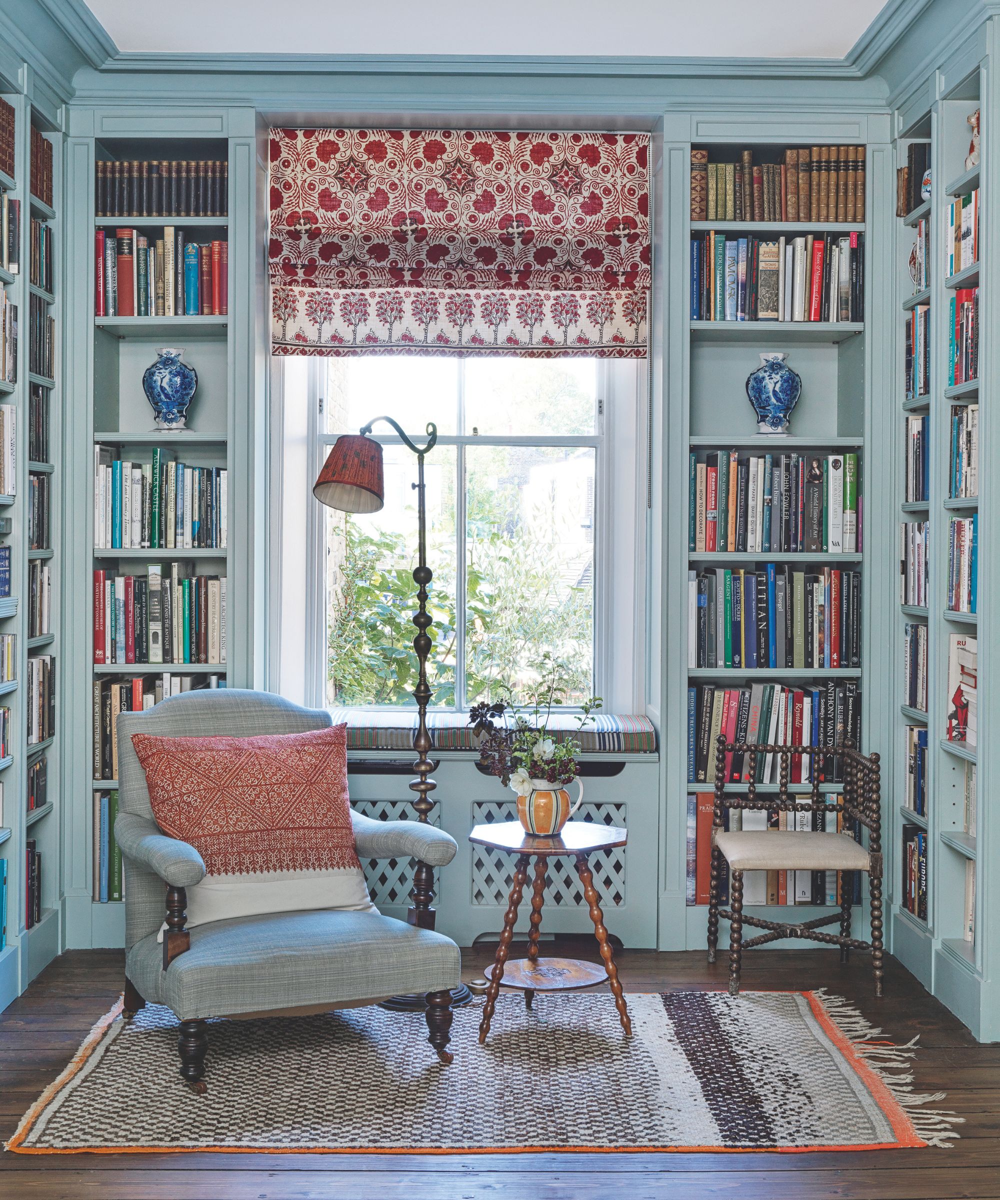 A blue living room with bookshelves, a blue upholstered chair, wooden floor lamp, and small wooden circular table. Behind is a large window with a red and white patterned blind.