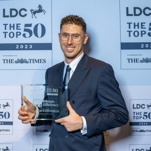 male wearing blue suit, white shirt and tie with short curly hair, wearing glasses, stood holding a glass award plaque