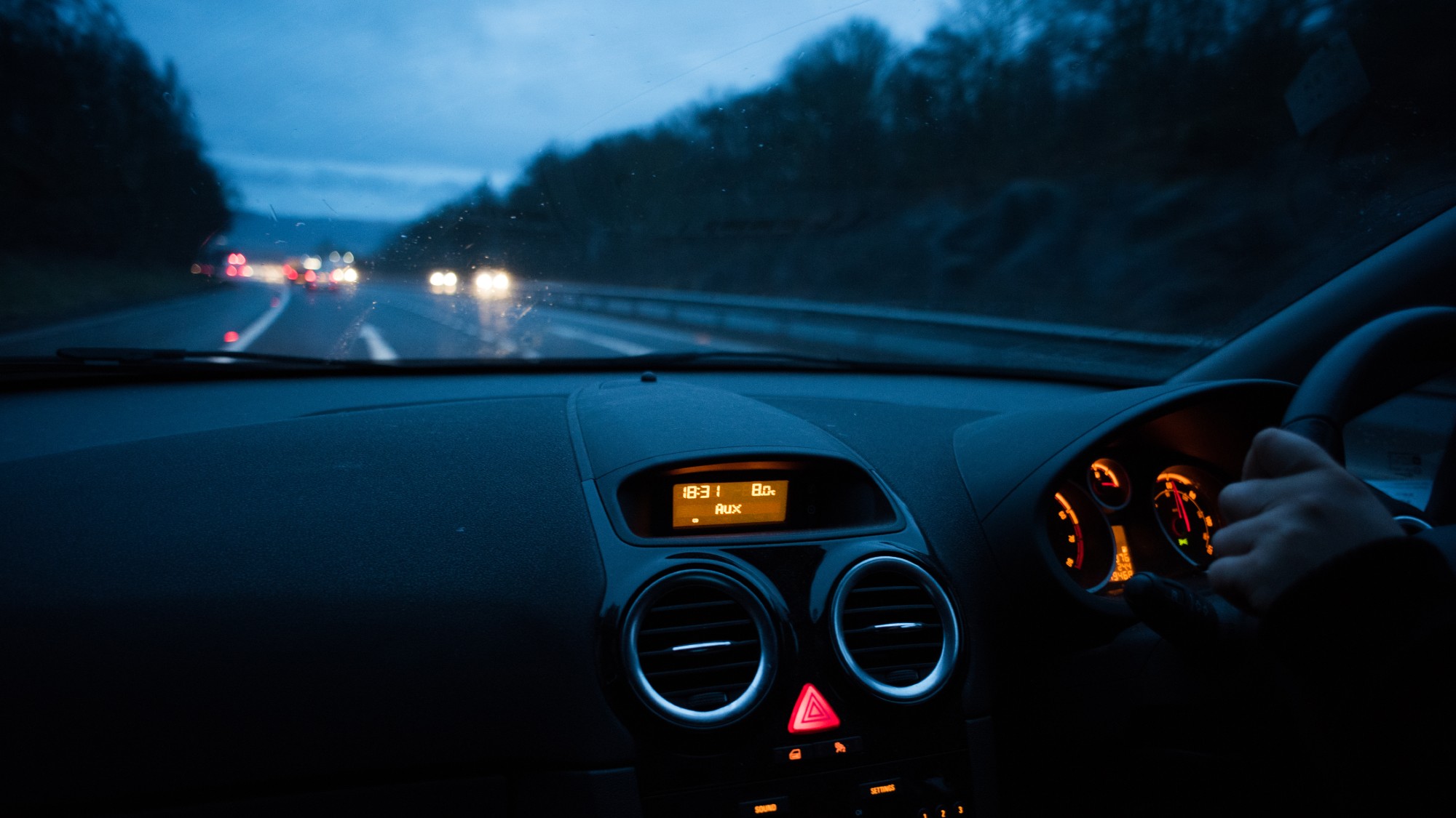 A UK road at night as viewed from the passenger side of the interior of a car, a hand grasping the steering wheel.