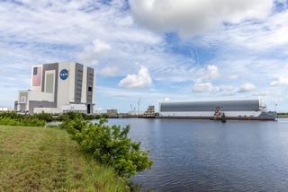 a square-shaped building with an american flag and nasa logo seen from far away. also visible is water with a long, low barge on it