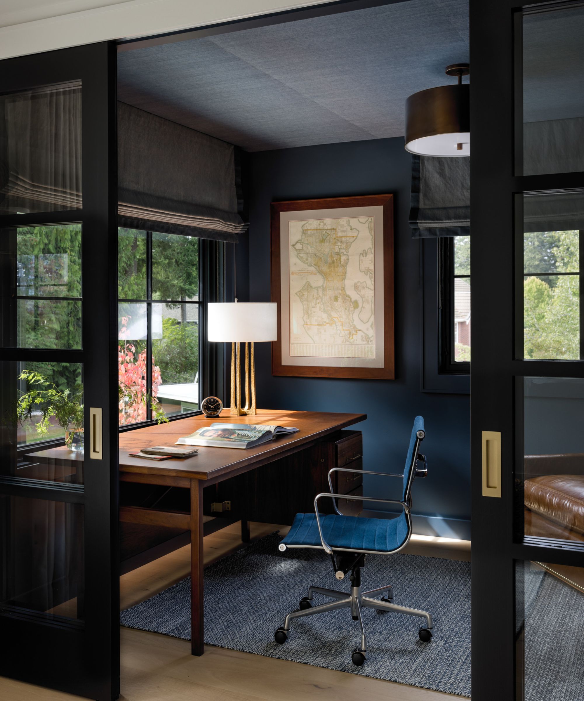 A small home office with dark navy walls, a blue sisal wallcovering on the ceiling, a large wooden desk, and a blue swivel chair.
