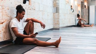 Man in shorts and T-shirt sitting on exercise mat, leaning against wall and looking at phone