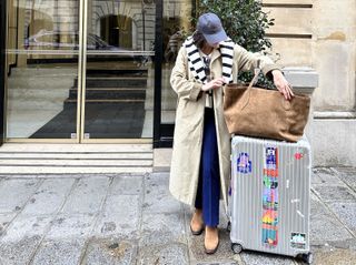 a woman standing outside a building next to her suitcase and tote wearing a baseball cap, blue jeans, and a tan trench coat with a striped sweater draped over her shoulders