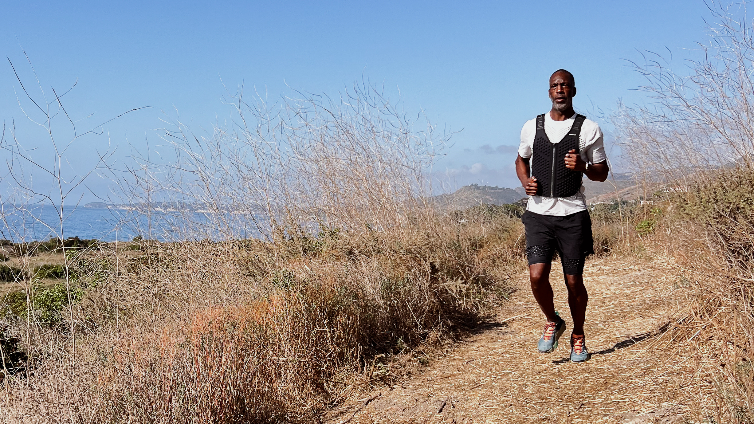 Man runs along trail wearing a black weighted vest with a view of the sea in the background