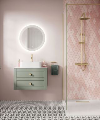 A small pink bathroom with light pink walls, a circular mirror, a sage green floating vanity with a white basin, a pink tiled shower with gold trim glass doors, and black and white flooring
