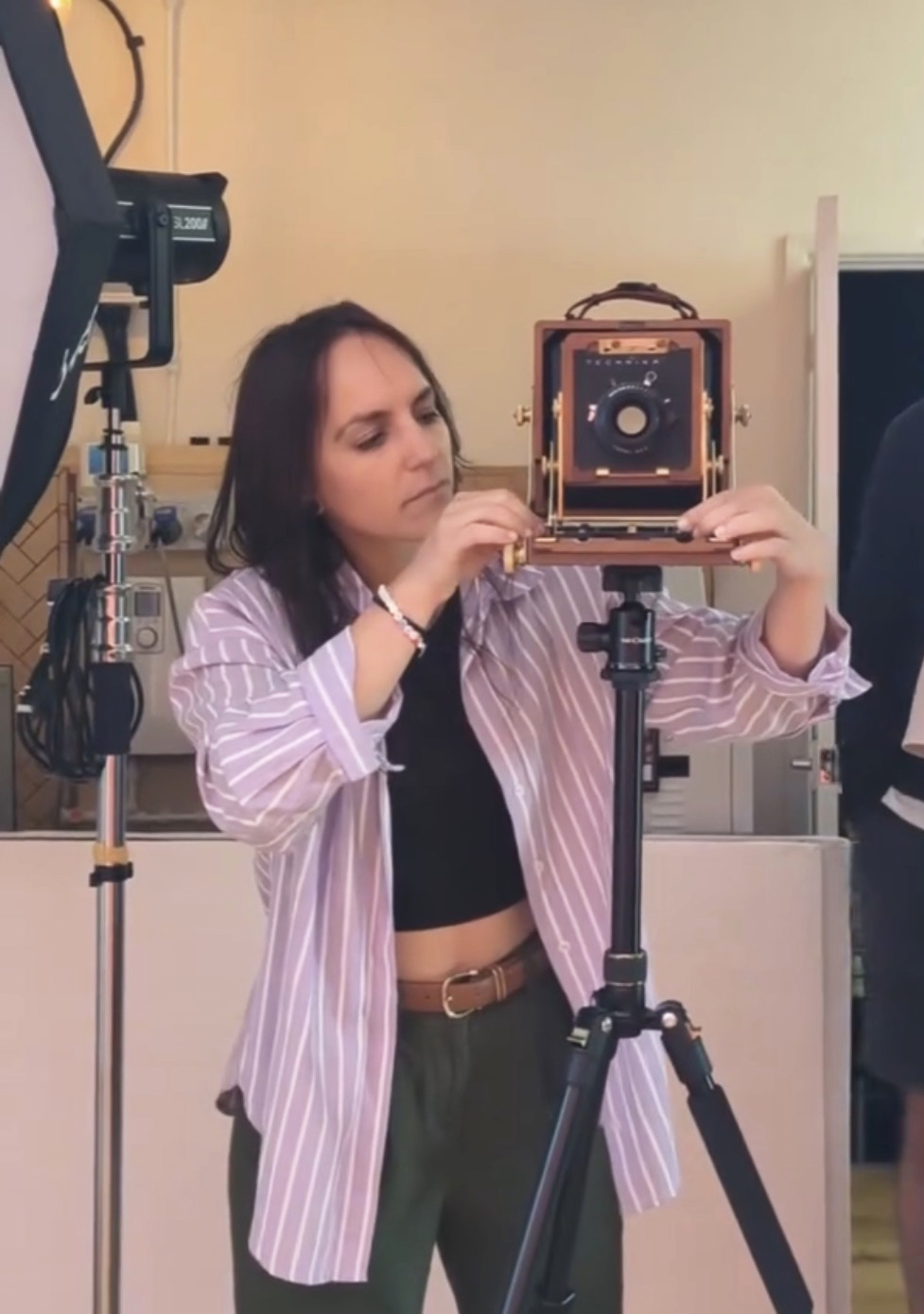 A person holds a vintage camera on a tripod, dressed in a striped shirt and green pants, in a modern photography studio