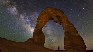 a person standing underneath the delicate arch in arches national park watching the milky way.