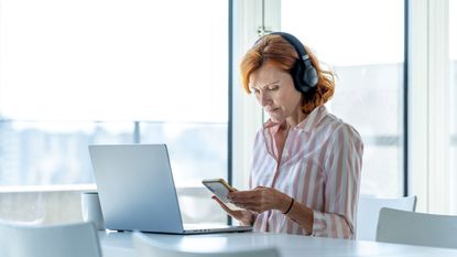 A woman looks with concern at her phone while listening to music on headphones sitting at a laptop. 