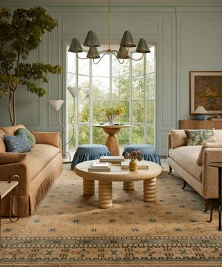 lulu and georgia spring living room painted pale blue with a large pastel rug and a caramel toned pleated sofa, sculptural coffee table, and blue striped ruffle ottomans