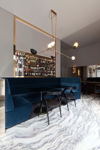 View of the interior of the restaurant with deep velvet blue and beige sitting area, a bar, gray and white marble flooring, and a huge spotlight chandelier.