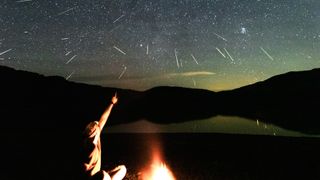  A figure sitting next to a campfire points up at a starry sky, streaked with shooting stars.