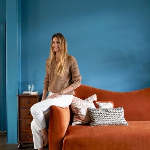 Image of a woman in white pants and a tan sweater sitting on the arm of an orange sofa in a blue room.