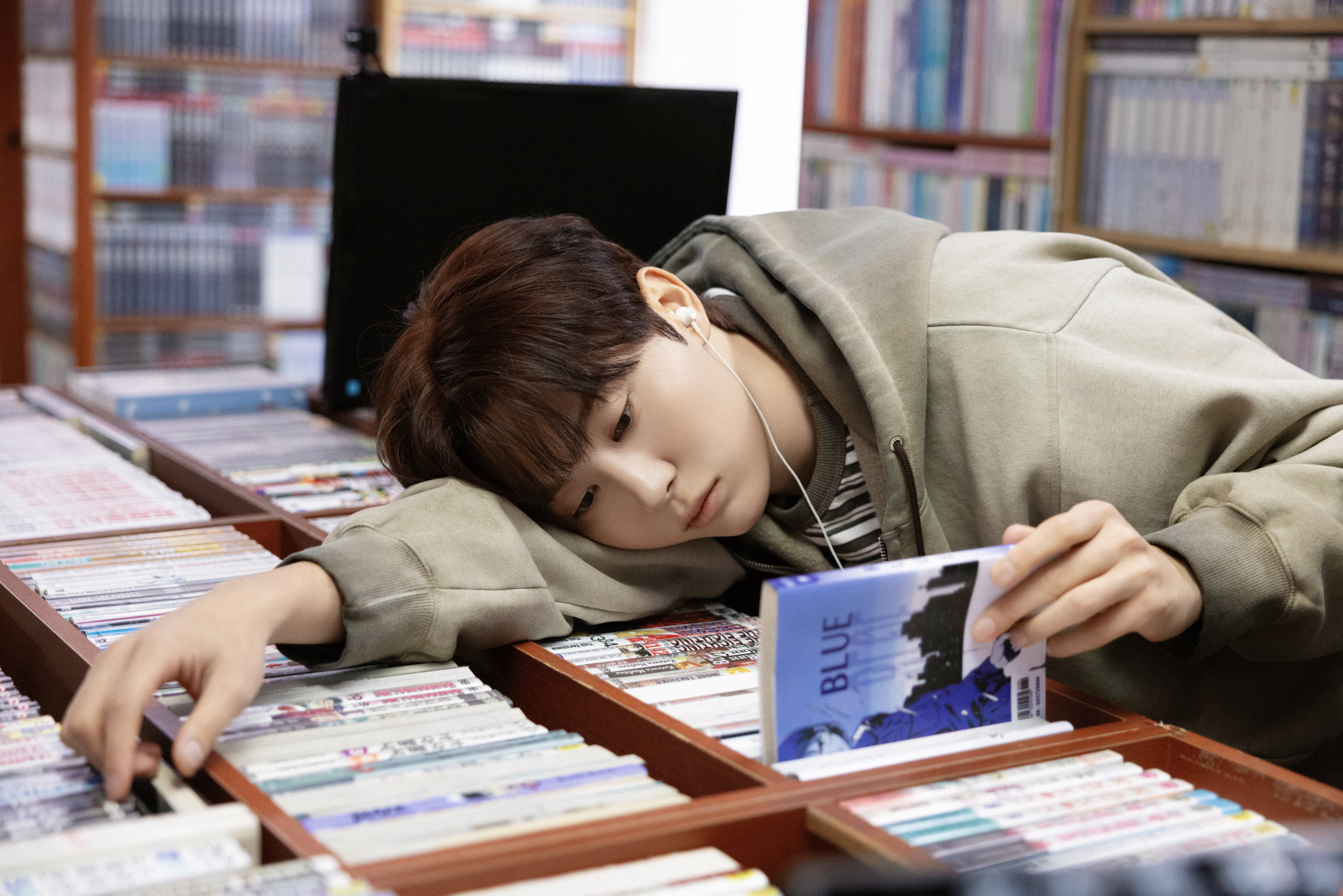 seungkwan from the k-pop group seventeen lies on a comic book display while wearing headphones and holding a book that reads "blue"