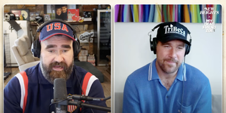 Travis and Jason Kelce, both wearing blue polos and baseball caps, speaking into mics on the New Heights Podcast