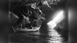 Two men stand in a rowing boat inside a sea cave, with daylight streaming through a rocky opening ahead of them.