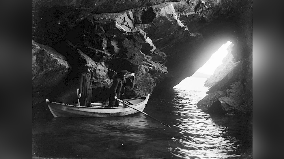 Two men stand in a rowing boat inside a sea cave, with daylight streaming through a rocky opening ahead of them.