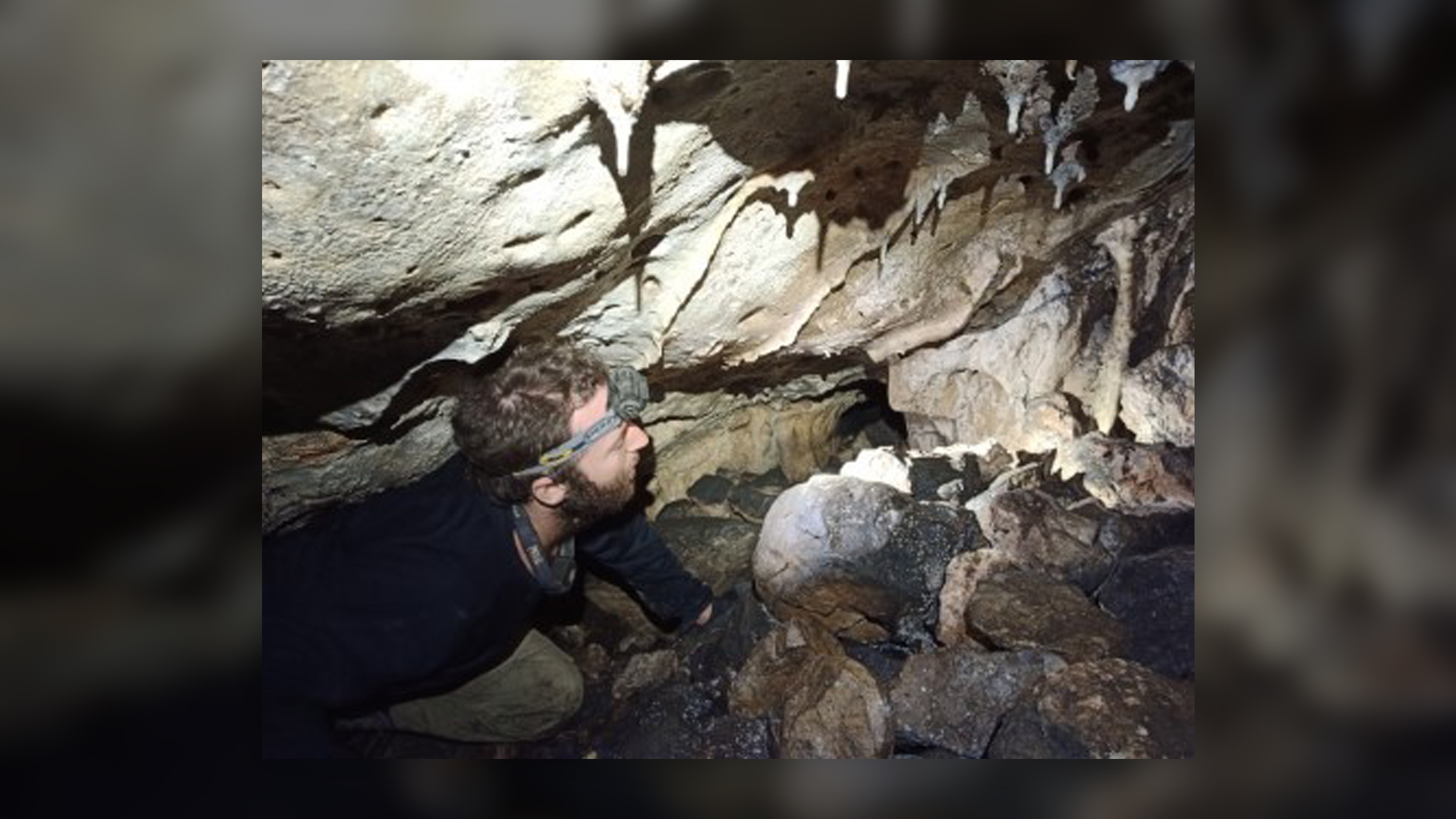 A man with a headlamp crawls on a rocky cave floor.